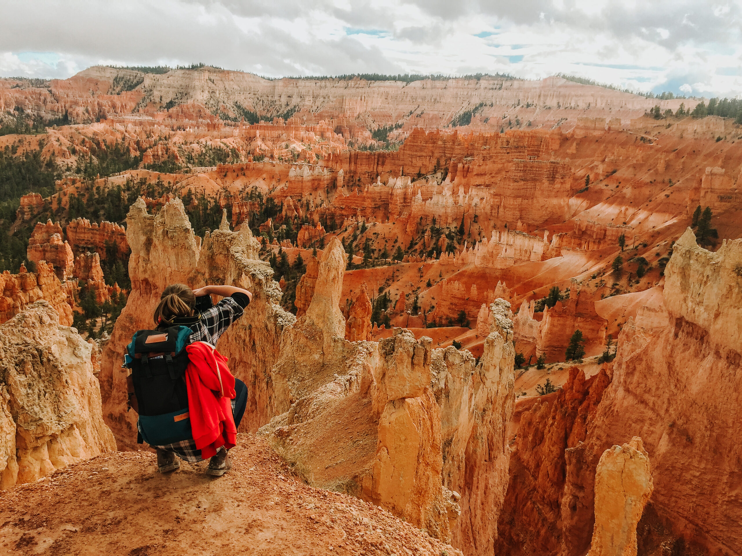 Krajobraz Wielkiego Kanionu Kolorado. Rude skały, niebo zachmurzone. Na pierwszym planie po lewej młoda dziewczyna z plecakiem, tyłem do widza, kuca i robi zdjęcie.