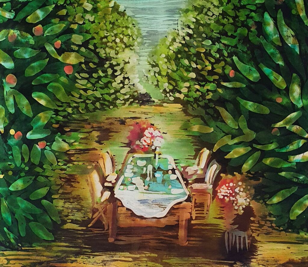 Zdjęcie pracy wykonanej metodą batiku. Na środku stół zastawiony do posiłku, otaczają go drzewa i krzewy.
